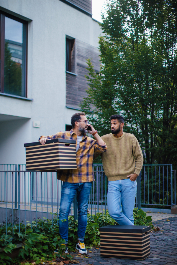 Man helping his friend move into new apartment in building. Man is moving into new flat, carrying box, waiting for moving truck and phone calling.