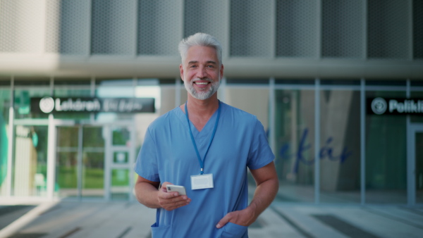 A handsome doctor looking at smartphone, standing in front of medical building or hospital, wearing blue uniform and ID card.