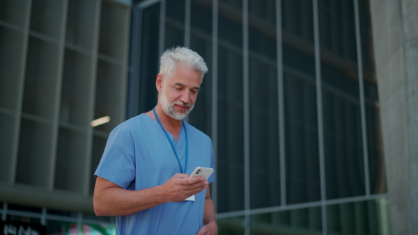 A handsome doctor looking at smartphone, standing in front of medical building or hospital, wearing blue uniform and ID card.