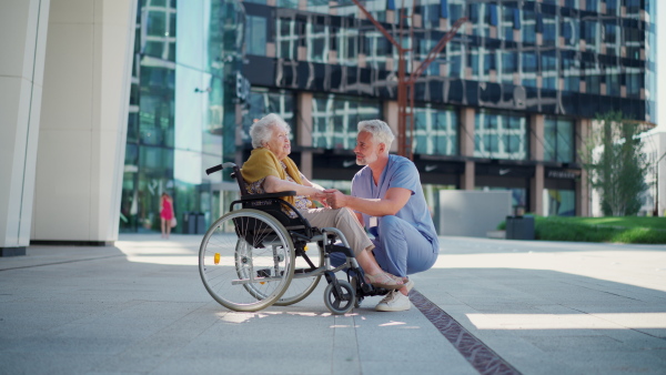 A male caregiver and elderly patient in wheelchair, holding hands. Geriatric nurse accompany lovely elderly woman on outdoor, looking at each other.