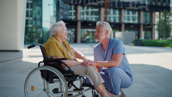 A male caregiver and elderly patient in wheelchair, holding hands. Geriatric nurse accompany lovely elderly woman on outdoor, looking at each other.