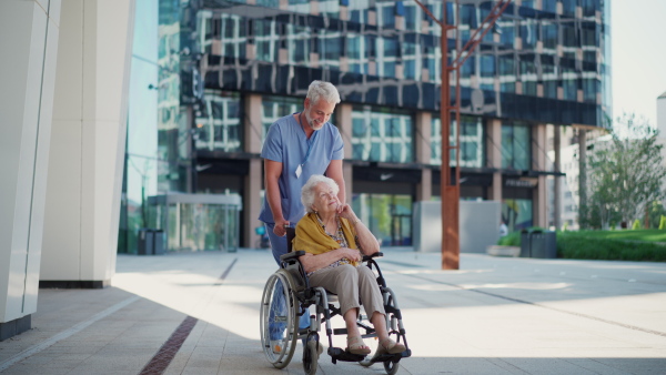 A male caregiver taking elderly patient on wheelchair stroll in the city. Geriatric nurse pushing wheelchair with lovely elderly woman.