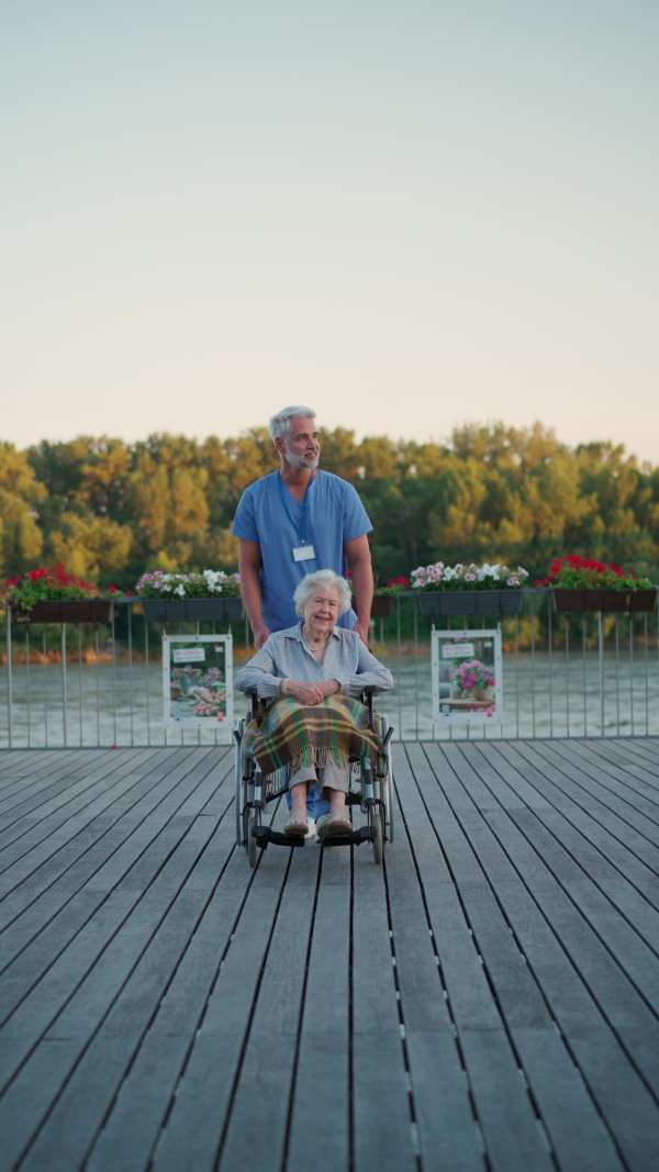 A male caregiver with elderly patient on wheelchair. Geriatric nurse accompany lovely elderly woman on outdoor walk, looking at river.