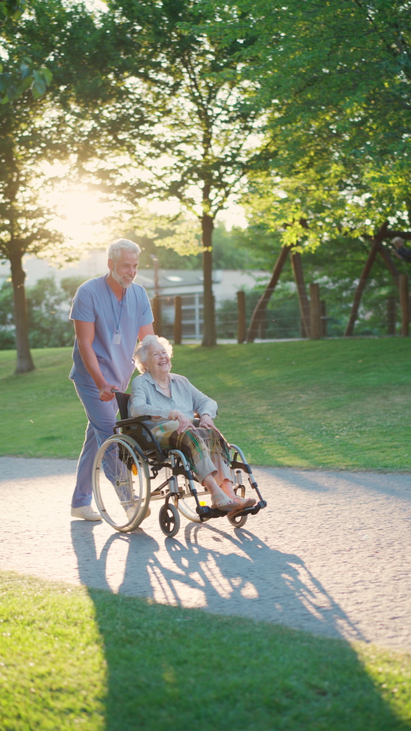 A video of male caregiver with elderly patient on wheelchair on stroll in the city park. Geriatric nurse taking senior in wheelchair on walk, enjoying warm autumn day.