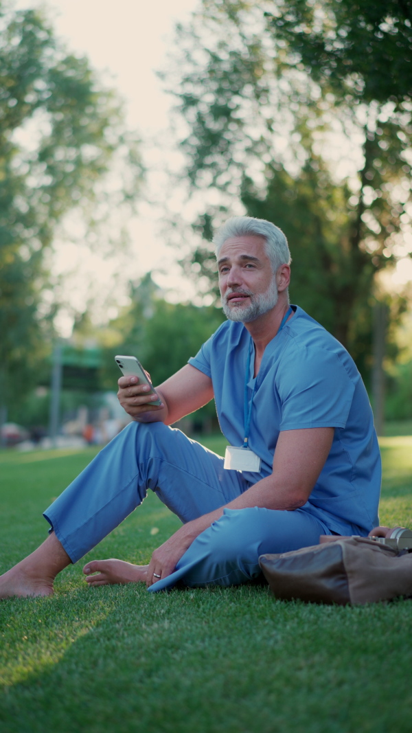 A handsome doctor in scrubs sitting on grass, relaxing with phone after long day in work. Job satisfaction, feeling contentment with his job. Mental health of healthcare worker.