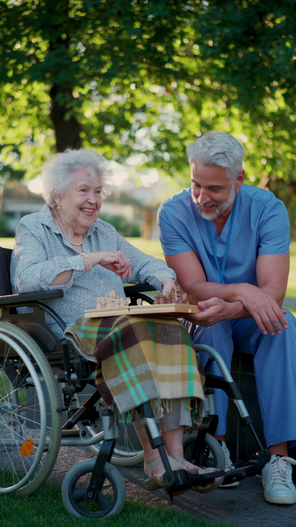 A senior woman in wheelchair playing chess with male caregiver. Geriatric nurse accompany lovely elderly patient in park, enjoying warm summer evening.