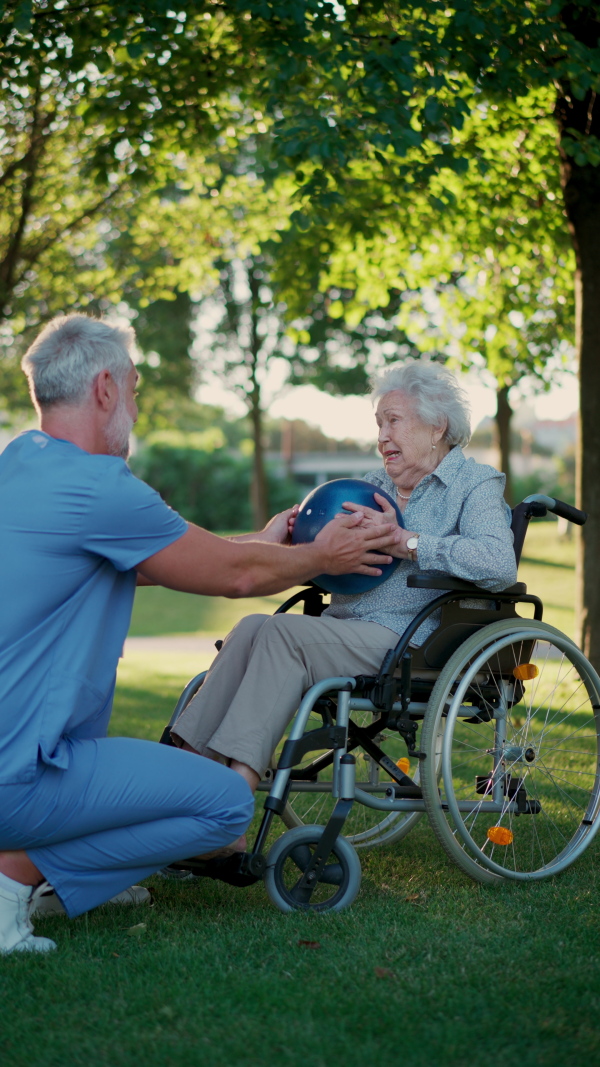 A male caregiver exercising with elderly patient on wheelchair, using small exercise ball. Geriatric physiotherapist during outdoor workout therapy in public park, nursing home garden.