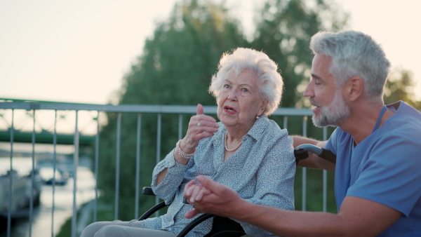 A male caregiver with elderly patient on wheelchair. Geriatric nurse accompany lovely elderly woman on outdoor walk, looking at river.