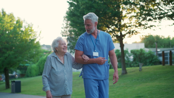 A male caregiver with elderly patient in park. Geriatric nurse accompany lovely elderly woman on outdoor walk, enjoying conversation.