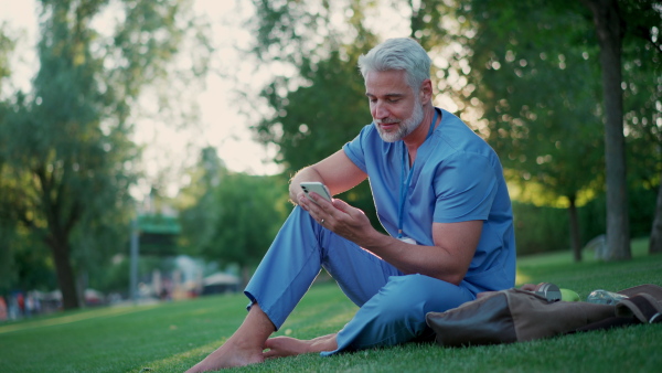 A handsome doctor in scrubs sitting on grass, relaxing with phone after long day in work. Job satisfaction, feeling contentment with his job. Mental health of healthcare worker.