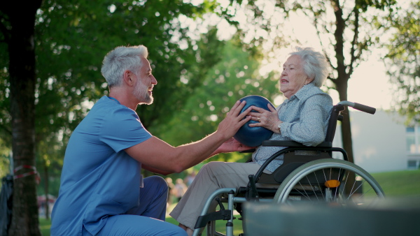 A male caregiver exercising with elderly patient on wheelchair, using small exercise ball. Geriatric physiotherapist during outdoor workout therapy in public park, nursing home garden.