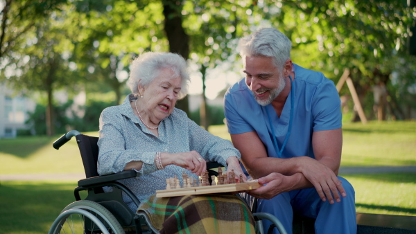 A senior woman in wheelchair playing chess with male caregiver. Geriatric nurse accompany lovely elderly patient in park, enjoying warm summer evening.