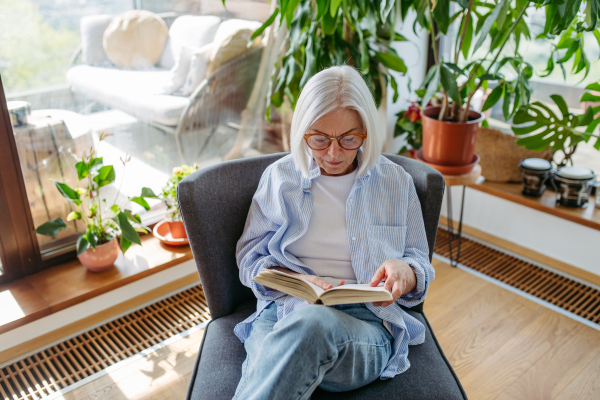 Beautiful mature woman reading book, feeling cozy and happy. Wearing reading glasses. Weekend activity for older woman, relaxing at home.