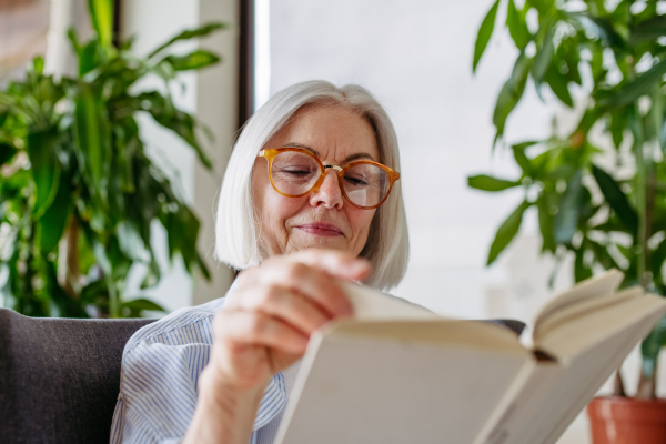 Beautiful mature woman reading book, feeling cozy and happy. Wearing reading glasses. Weekend activity for older woman, relaxing at home.