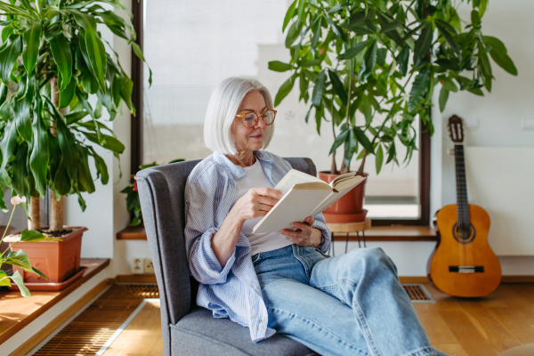 Beautiful mature woman reading book, feeling cozy and happy. Wearing reading glasses. Weekend activity for older woman, relaxing at home.