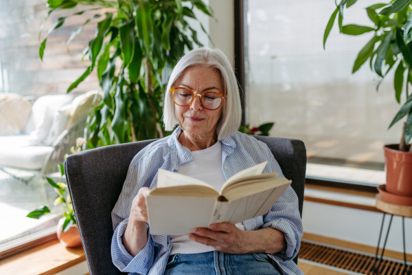 Beautiful mature woman reading book, feeling cozy and happy. Wearing reading glasses. Weekend activity for older woman, relaxing at home.