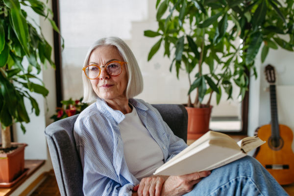 Beautiful mature woman reading book, feeling cozy and happy. Wearing reading glasses. Weekend activity for older woman, relaxing at home.