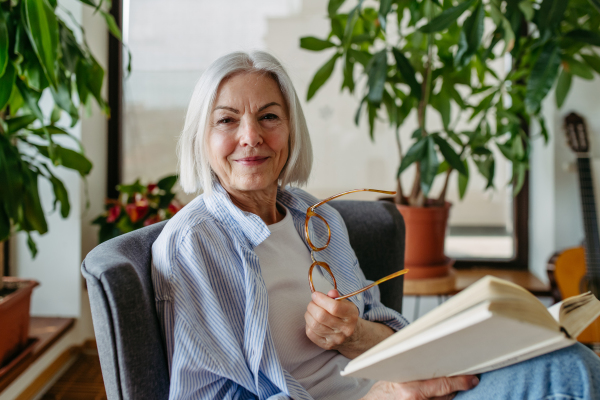 Beautiful mature woman reading book, feeling cozy and happy. Wearing reading glasses. Weekend activity for older woman, relaxing at home.