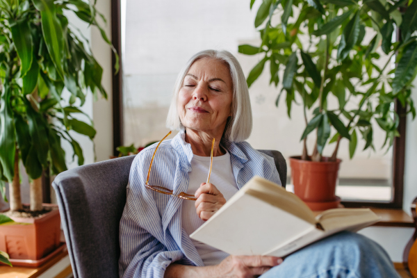 Beautiful mature woman reading book, feeling cozy and happy. Wearing reading glasses. Weekend activity for older woman, relaxing at home.