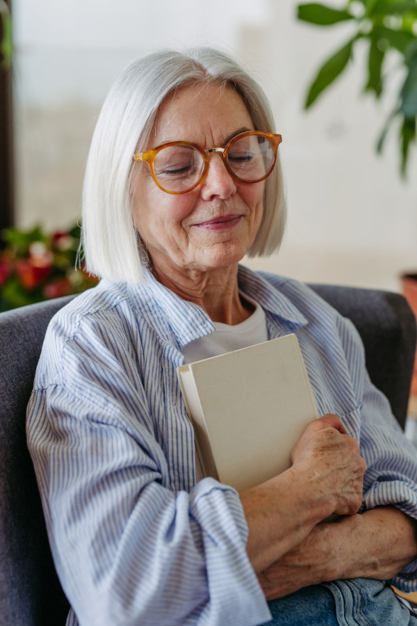 Beautiful mature woman reading book, feeling cozy and happy. Wearing reading glasses. Weekend activity for older woman, relaxing at home.