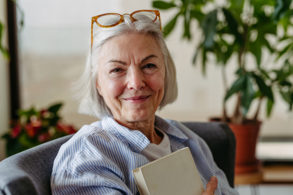 Beautiful mature woman reading book, feeling cozy and happy. Wearing reading glasses. Weekend activity for older woman, relaxing at home.