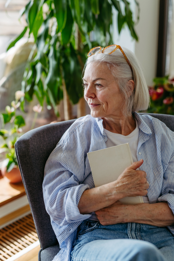 Beautiful mature woman reading book, feeling cozy and happy. Wearing reading glasses. Weekend activity for older woman, relaxing at home.