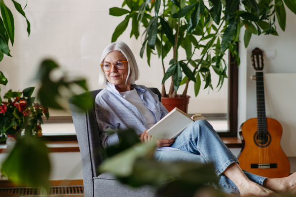 Beautiful mature woman reading book, feeling cozy and happy. Wearing reading glasses. Weekend activity for older woman, relaxing at home.