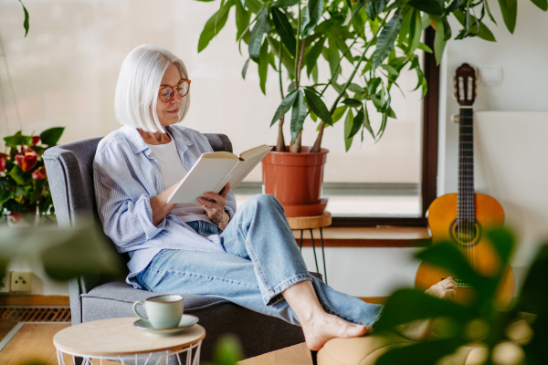 Beautiful mature woman reading book, feeling cozy and happy. Wearing reading glasses. Weekend activity for older woman, relaxing at home.