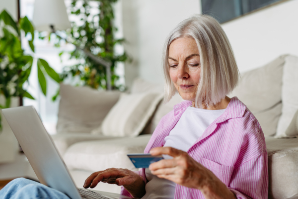 Mature woman paying with debit, credit card online, using internet banking and payment gateway. Shopping online, paying bills and risk of frauds and hoax. Attractive woman sitting on floor.