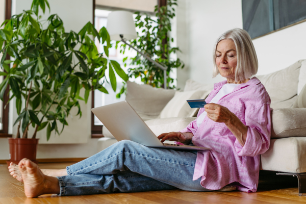 Mature woman paying with debit, credit card online, using internet banking and payment gateway. Shopping online, paying bills and risk of frauds and hoax. Attractive woman sitting on floor.