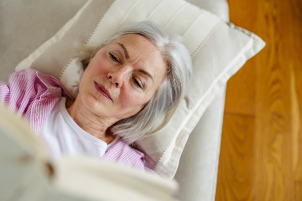 Beautiful mature woman reading book, feeling cozy and happy. Wearing reading glasses. Weekend activity for older woman, relaxing at home.