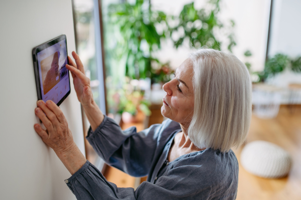 Mature woman adjusting smart thermostat, touch screen of smart home device. Older woman using smart technology at her home.