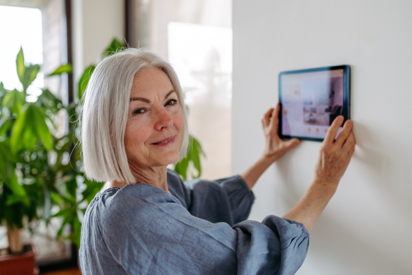 Mature woman adjusting smart thermostat, touch screen of smart home device. Older woman using smart technology at her home.