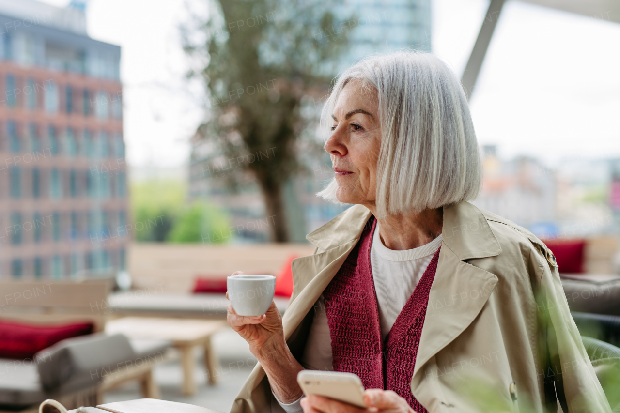 Attractive older woman checking smartphone, drinking coffee at coffee shop. Mature woman spending free time outdoors, in cafe, waiting for her girl friends.