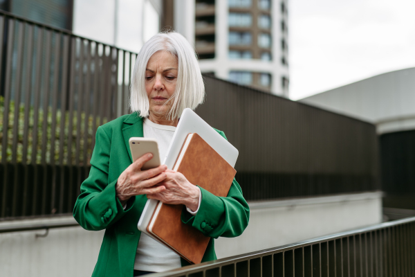 Mature businesswoman waiting on bus after wok, scrolling on smartphone. Beautiful older woman with gray hair standing on city street, waiting on bus.