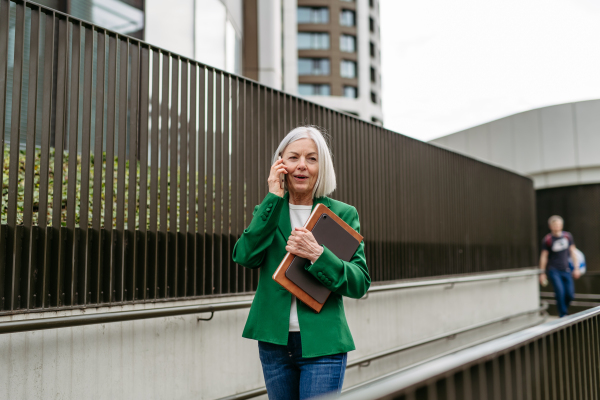 Mature businesswoman phone calling smartphone, going on business meeting in the city. Beautiful older woman with gray hair standing on city street, waiting on bus.