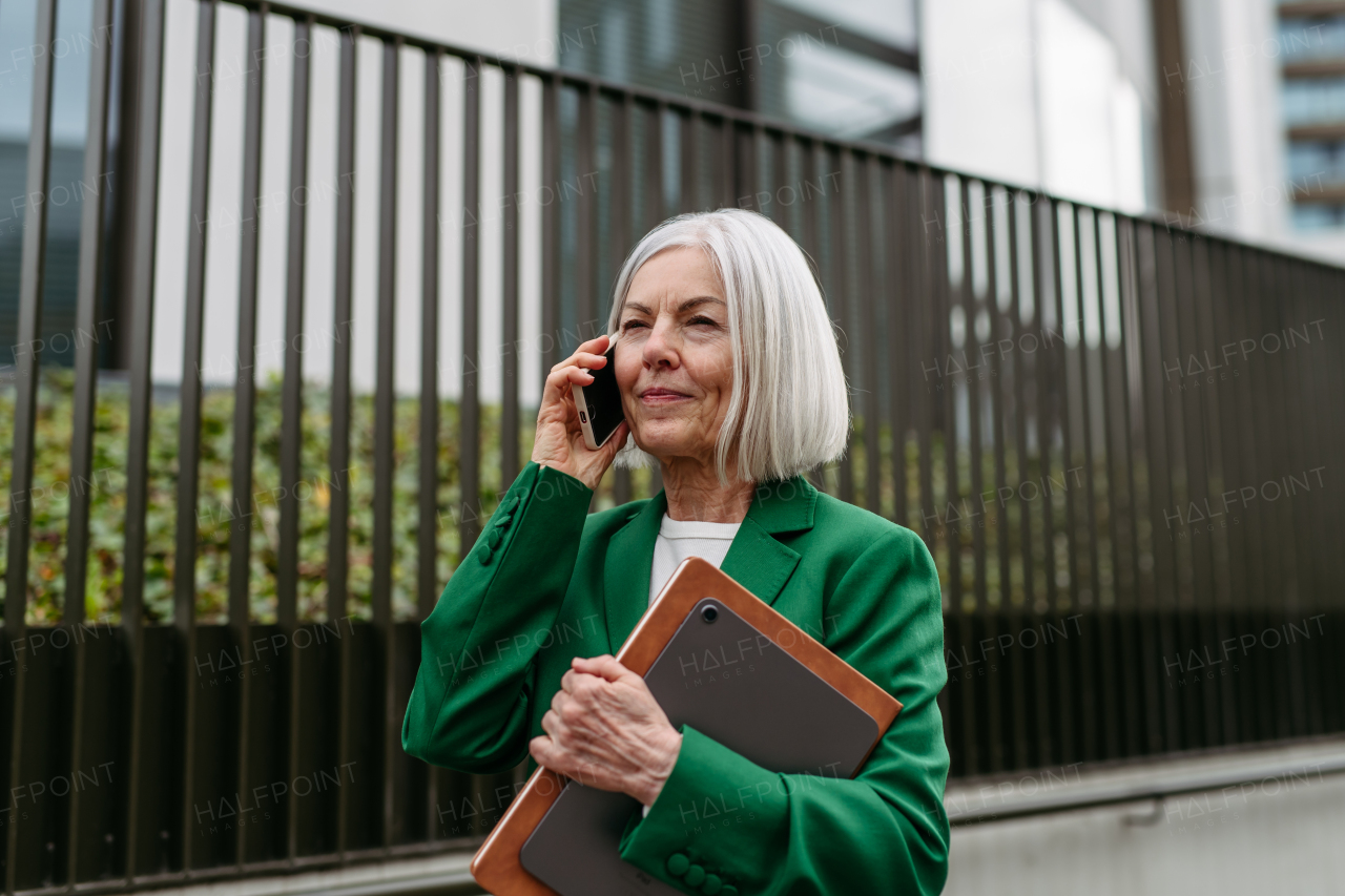 Mature businesswoman phone calling smartphone, going on business meeting in the city. Beautiful older woman with gray hair standing on city street, waiting on bus.