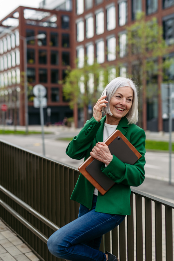 Mature businesswoman phone calling smartphone, going on business meeting in the city. Beautiful older woman with gray hair standing on city street, waiting on bus.