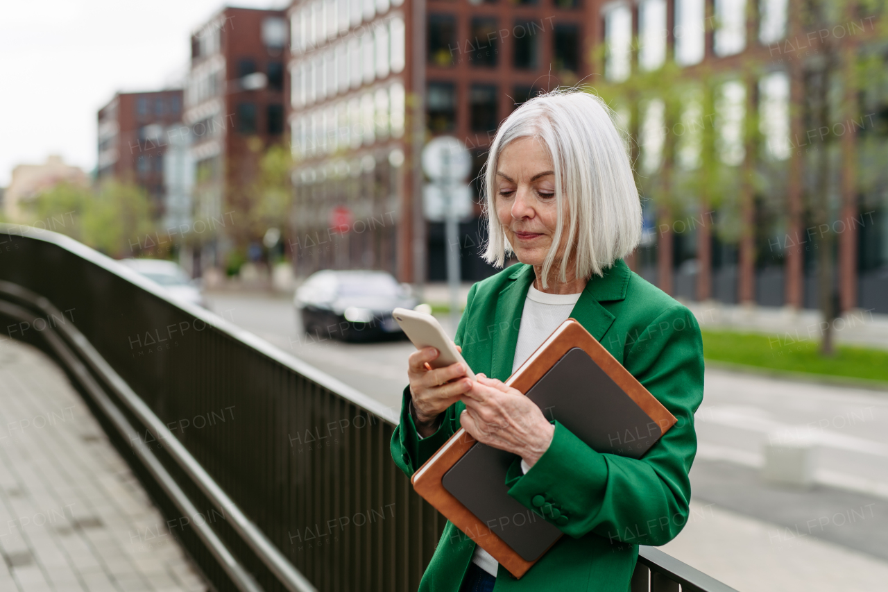 Mature businesswoman scrolling on smartphone, going home from work. Beautiful older woman with gray hair standing on city street, waiting on bus.