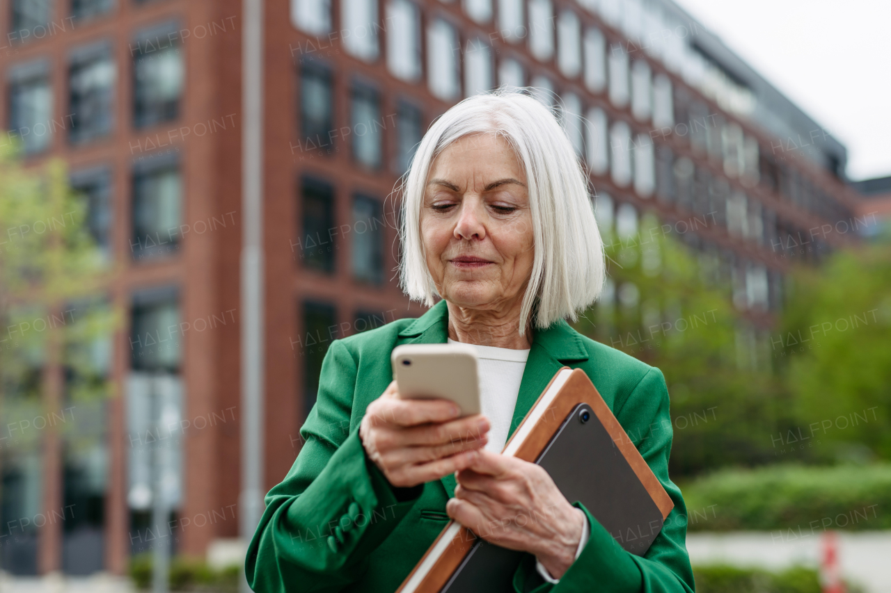 Mature businesswoman waiting on bus after wok, scrolling on smartphone. Beautiful older woman with gray hair standing on city street, waiting on bus.