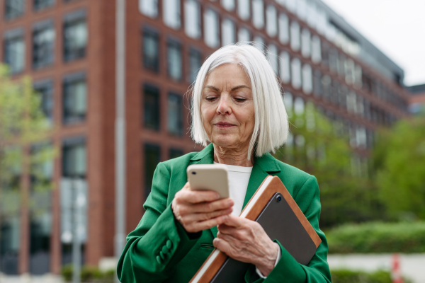 Mature businesswoman waiting on bus after wok, scrolling on smartphone. Beautiful older woman with gray hair standing on city street, waiting on bus.