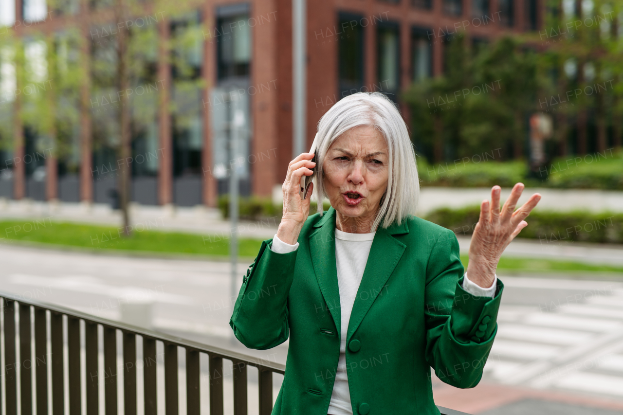 Mature businesswoman is angry, phone calling smartphone with business partner. Phone conflict, difficult customer . Beautiful older woman with gray hair standing on city street.