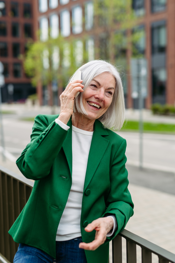 Mature businesswoman phone calling on smartphone, going on business meeting in the city. Beautiful older woman with gray hair standing on city street, smiling.