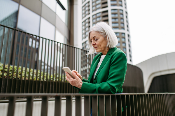 Mature businesswoman holding smartphone, waiting for business partner in the citBeautiful older woman with gray hair standing on city street, smiling.