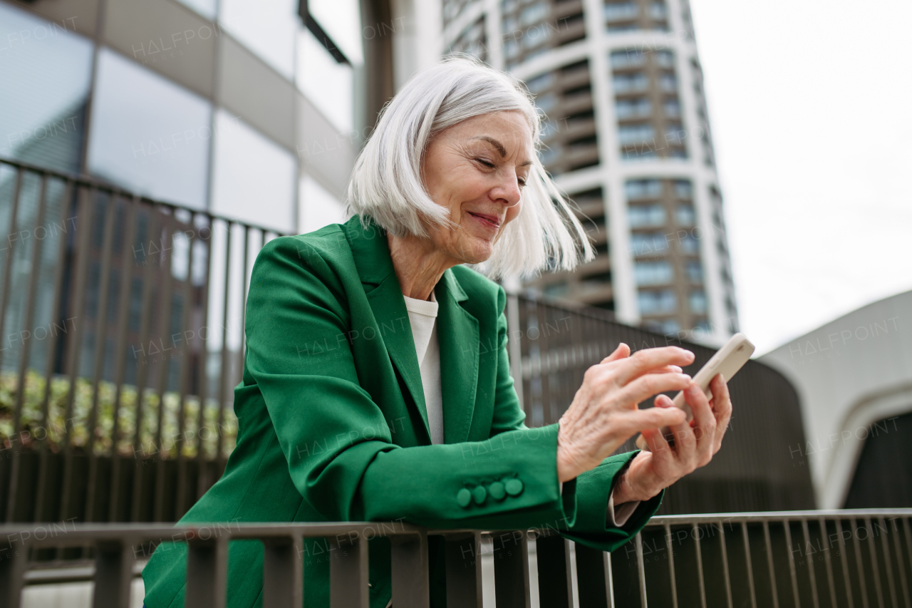 Mature businesswoman scrolling on smartphone, going home from work. Beautiful older woman with gray hair standing on city street, waiting on bus.