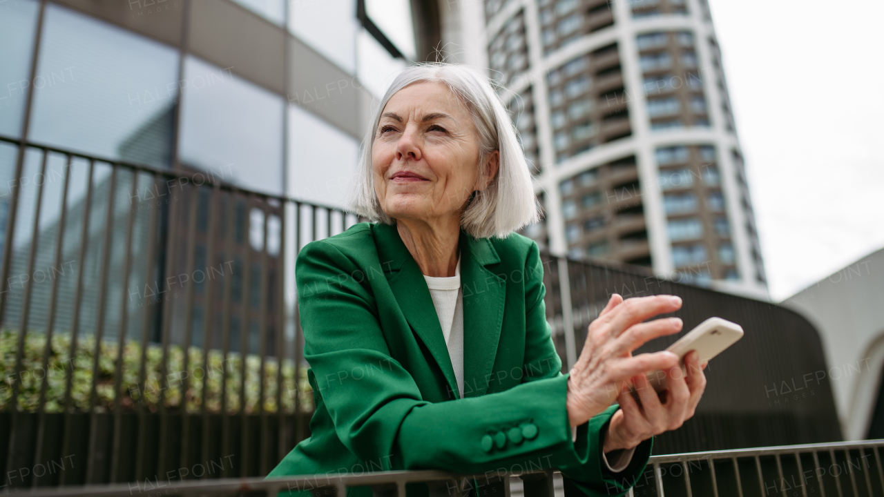 Mature businesswoman holding smartphone, waiting for business partner in the citBeautiful older woman with gray hair standing on city street, smiling.