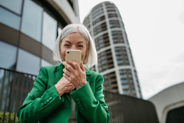 Mature woman holding smartphone in front of her face, going home from work. Beautiful older woman with gray hair standing on city street.