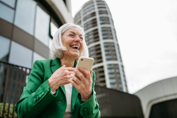 Mature businesswoman scrolling on smartphone, going home from work. Beautiful older woman with gray hair standing on city street, waiting on bus.