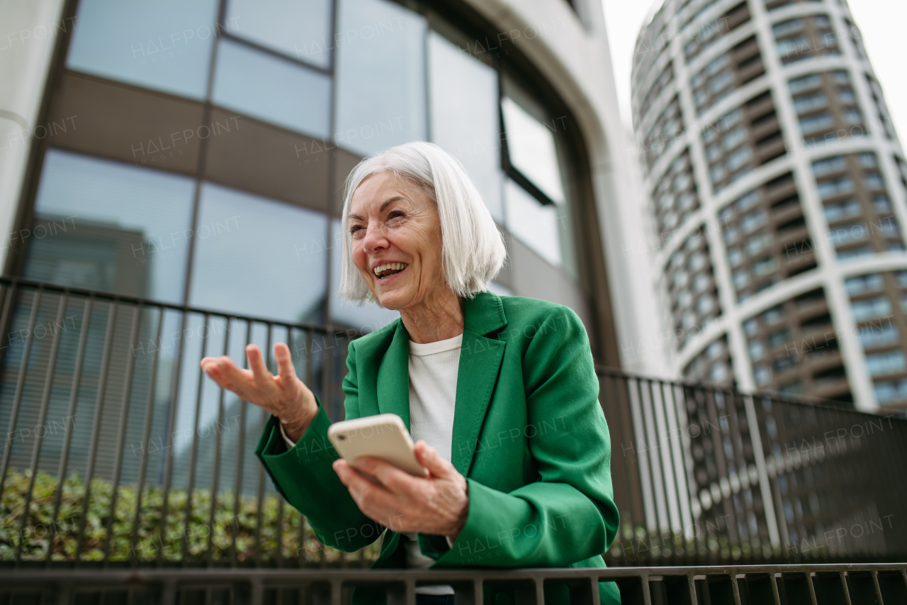 Mature businesswoman scrolling on smartphone, going home from work. Beautiful older woman with gray hair standing on city street, waiting on bus.