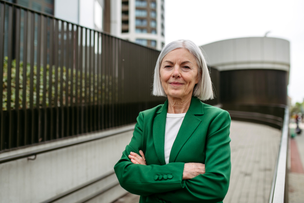 Portrait of mature businesswoman in front of office building. Beautiful older woman with gray hair, green jacket on city street.